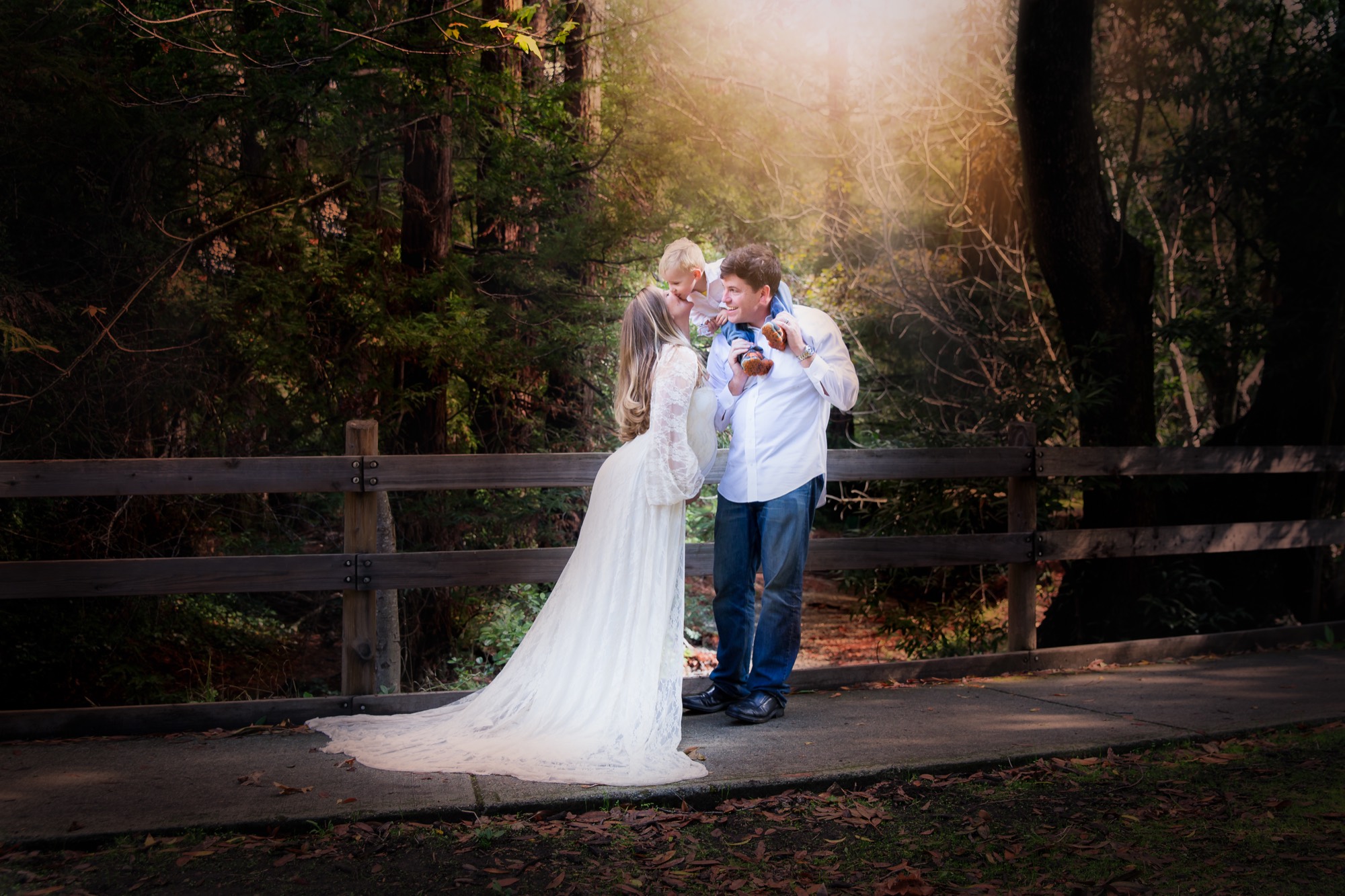 A family of three poses for a portrait on a paved path next to a wooden fence in a wooded park. The mother, wearing a long white lace dress, kisses her young child, who is sitting on the father's shoulders.