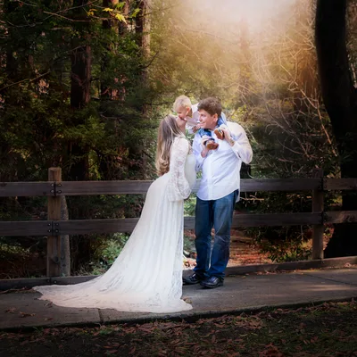 A family of three poses for a portrait on a paved path next to a wooden fence in a wooded park. The mother, wearing a long white lace dress, kisses her young child, who is sitting on the father's shoulders.