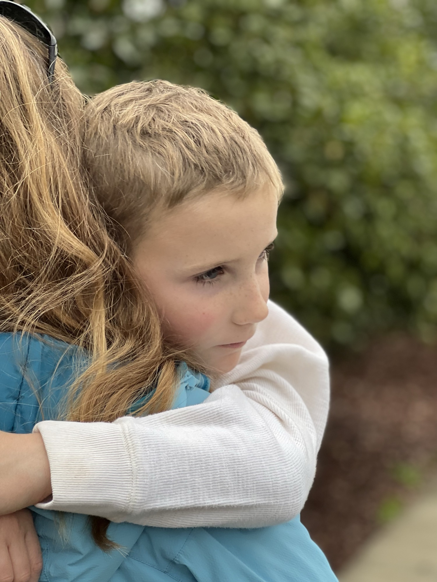 A close-up shot captures a young child with a pensive expression hugging a long-haired person wearing a blue jacket. The embrace takes place outdoors, with a soft-focus background of green foliage.