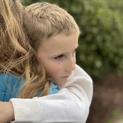 A close-up shot captures a young child with a pensive expression hugging a long-haired person wearing a blue jacket. The embrace takes place outdoors, with a soft-focus background of green foliage.
