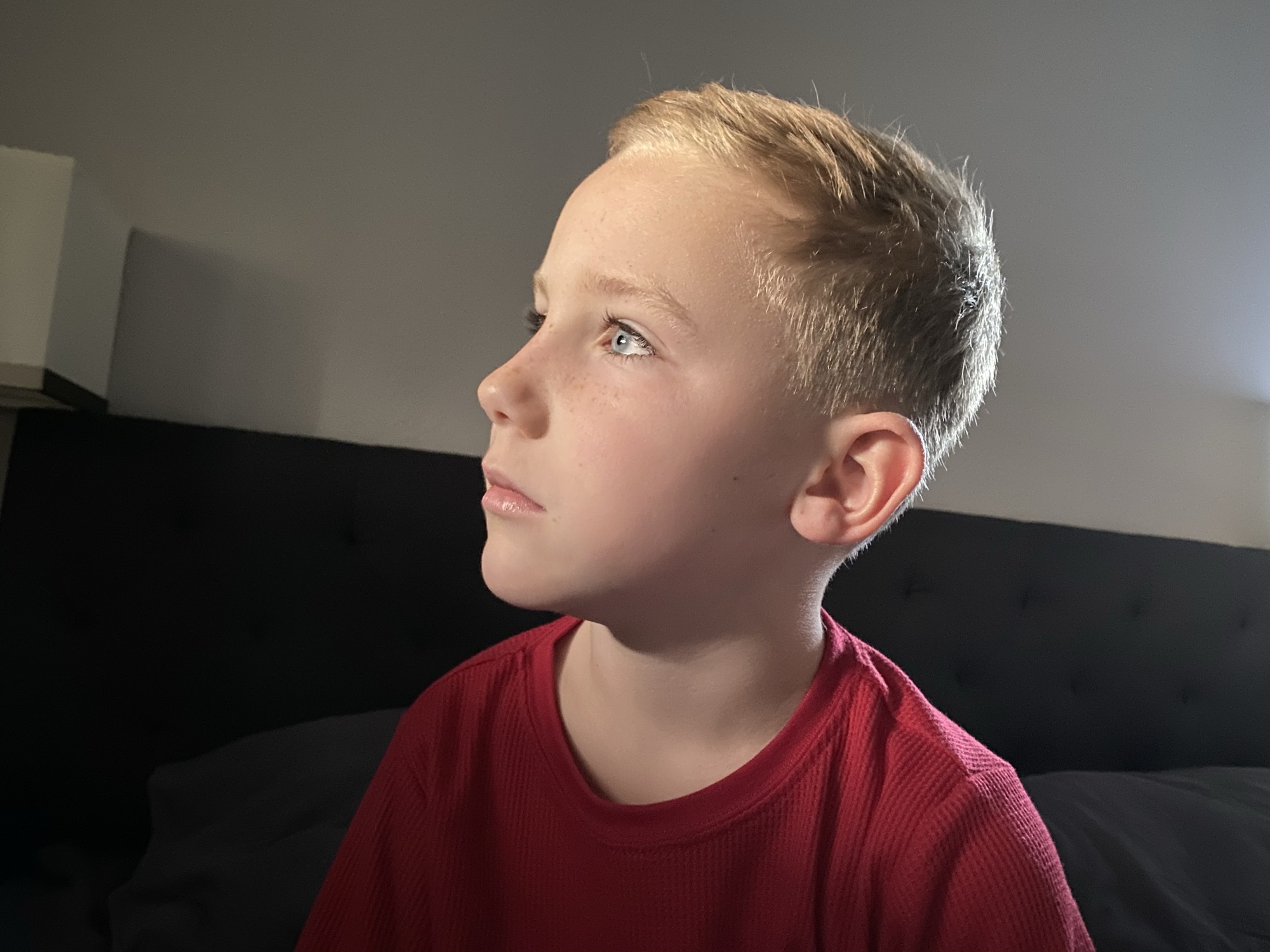 A close-up portrait of a young boy with short blonde hair and blue eyes looking off-camera in a dimly lit indoor setting. He is wearing a red shirt and is positioned in front of a dark headboard.