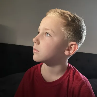 A close-up portrait of a young boy with short blonde hair and blue eyes looking off-camera in a dimly lit indoor setting. He is wearing a red shirt and is positioned in front of a dark headboard.