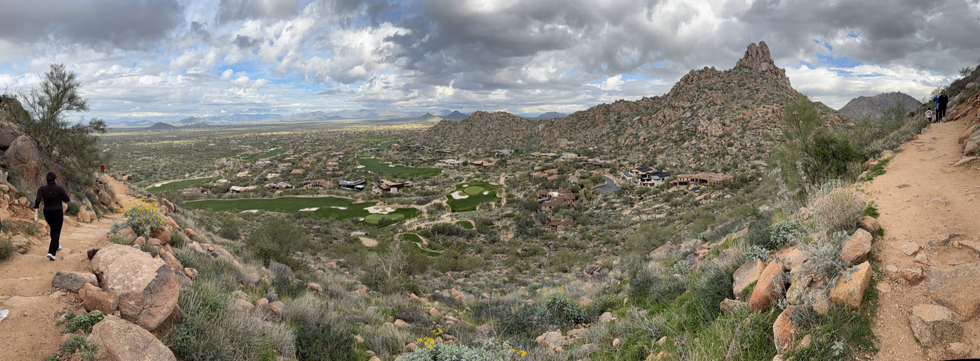 This panoramic photograph captures a wide view of a desert valley with a golf course and residential homes, taken from a rocky hiking trail on a cloudy day. Several hikers are visible on the dirt paths on either side of the frame, overlooking the expansive landscape below.