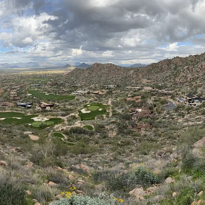 This panoramic photograph captures a wide view of a desert valley with a golf course and residential homes, taken from a rocky hiking trail on a cloudy day. Several hikers are visible on the dirt paths on either side of the frame, overlooking the expansive landscape below.