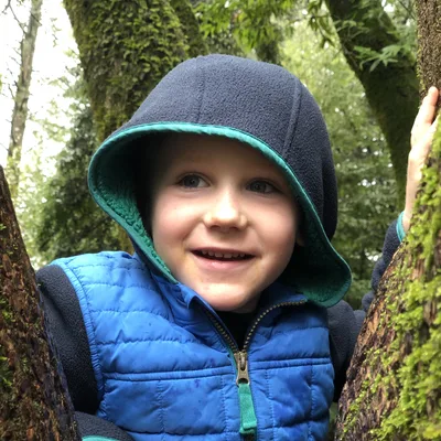 A young child wearing a blue hooded jacket smiles while peering out from between two moss-covered tree trunks in a forest.
