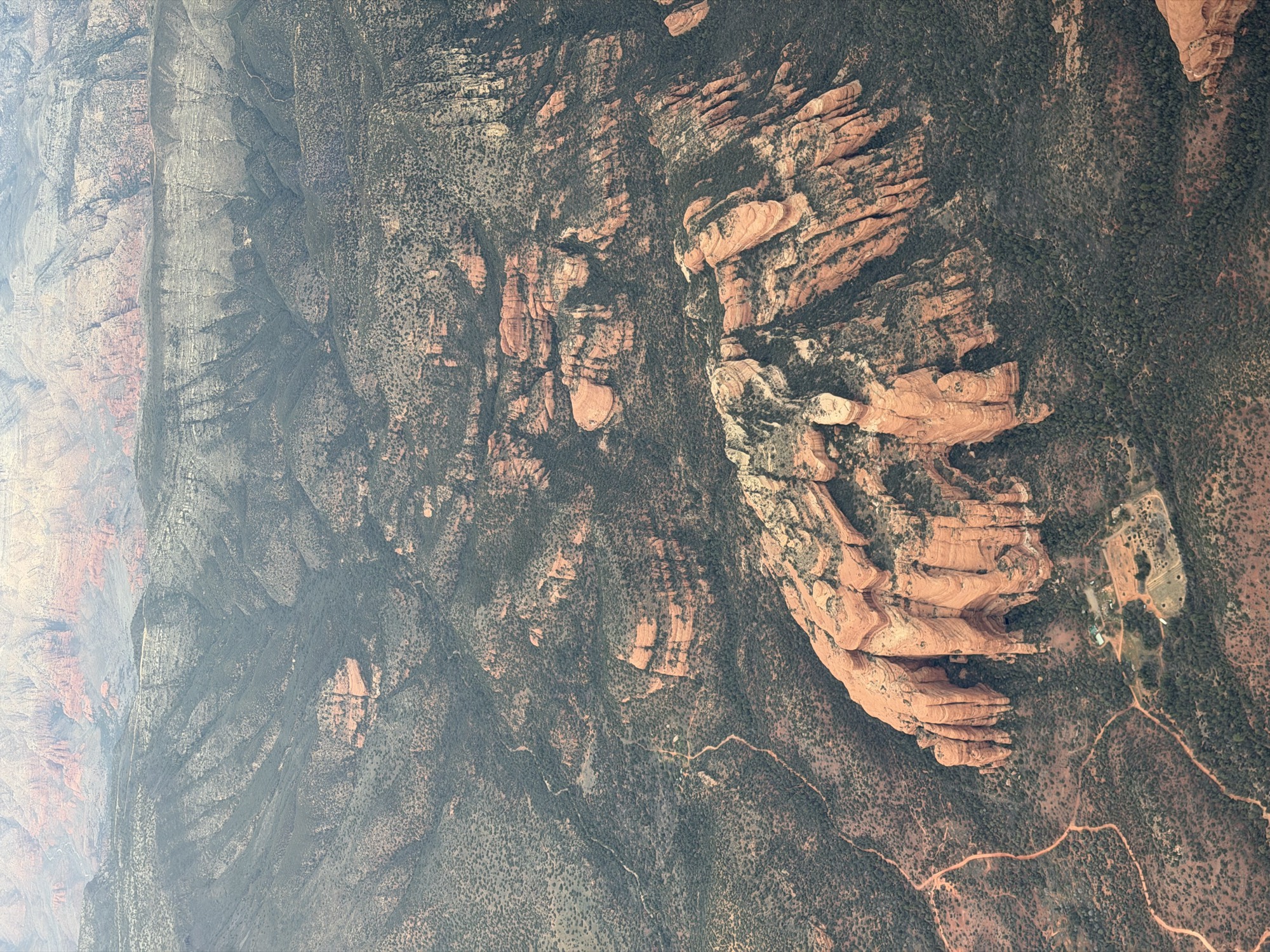An aerial view captures a vast desert landscape featuring prominent red rock formations, canyons, and sparse green vegetation. A winding dirt path and a small cluster of structures are visible near the base of the largest rock formation.