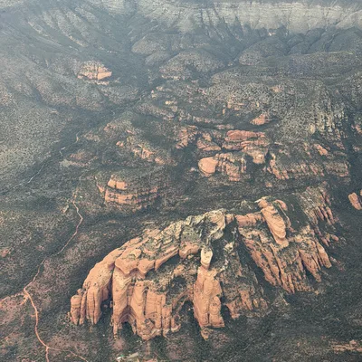 An aerial view captures a vast desert landscape featuring prominent red rock formations, canyons, and sparse green vegetation. A winding dirt path and a small cluster of structures are visible near the base of the largest rock formation.