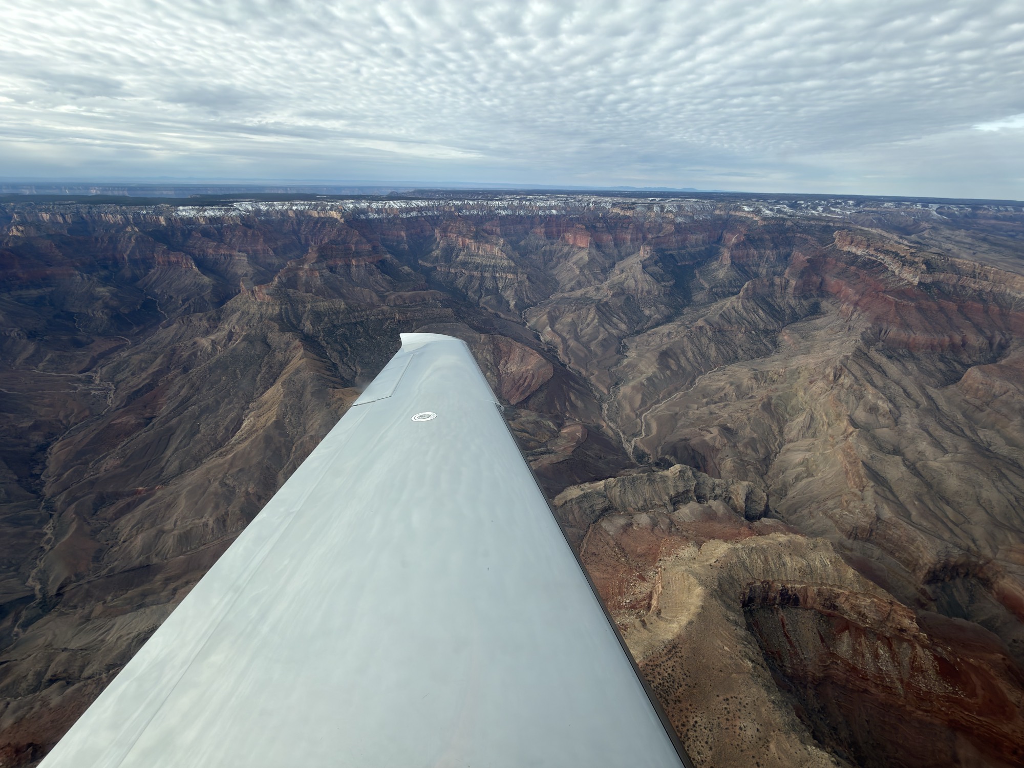 An aerial view taken from an airplane shows its wing extending over the vast, rugged expanse of the Grand Canyon under a cloudy sky. The distant canyon rims are lightly dusted with snow, highlighting the deep gorges and rock formations below.