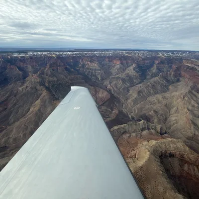 An aerial view taken from an airplane shows its wing extending over the vast, rugged expanse of the Grand Canyon under a cloudy sky. The distant canyon rims are lightly dusted with snow, highlighting the deep gorges and rock formations below.