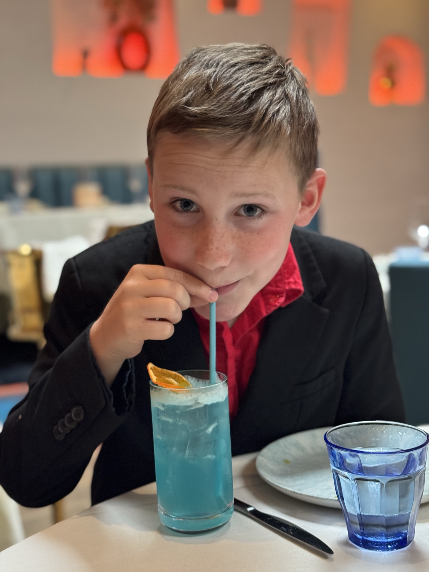 A young boy wearing a black suit jacket and red shirt sits at a restaurant table, looking at the camera while sipping a blue iced drink through a straw. The table also holds a plate, a knife, and a blue glass of water, with a softly blurred background showing warm lighting.