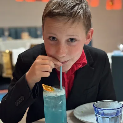 A young boy wearing a black suit jacket and red shirt sits at a restaurant table, looking at the camera while sipping a blue iced drink through a straw. The table also holds a plate, a knife, and a blue glass of water, with a softly blurred background showing warm lighting.