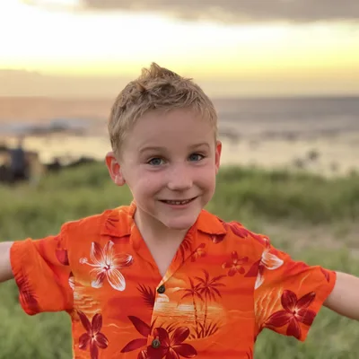 A young boy wearing an orange Hawaiian shirt smiles with his arms outstretched in a grassy area. In the background, the ocean is visible under a sunset sky.