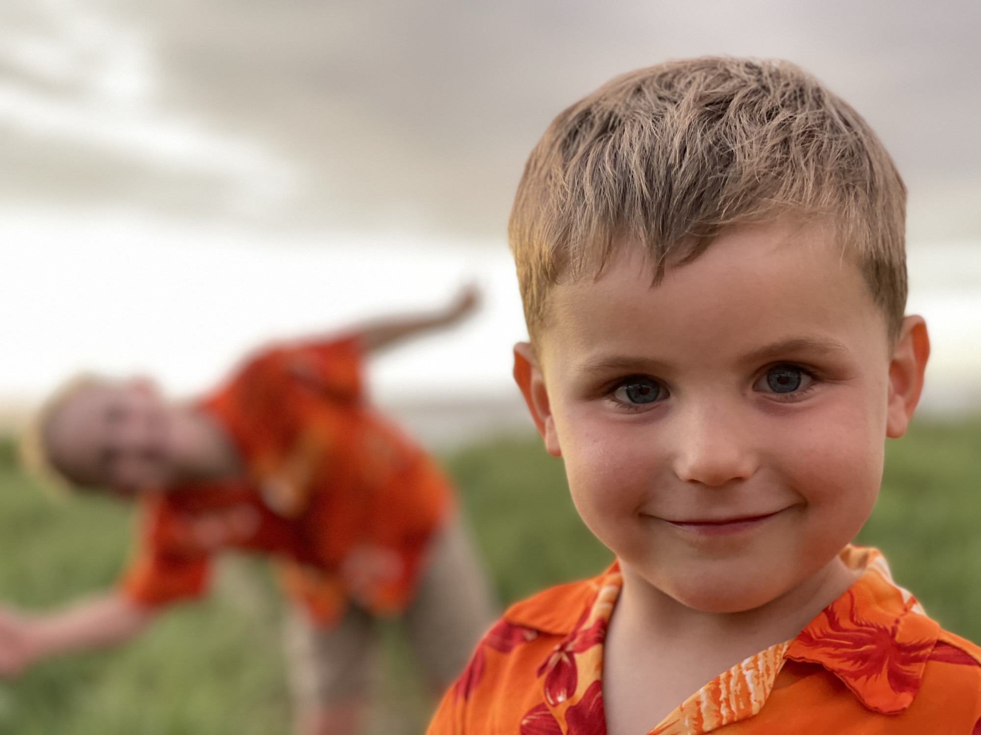A young boy in an orange floral shirt smiles at the camera in a close-up portrait, while another child in matching attire is blurred in the background, posing playfully in an outdoor grassy field under an overcast sky.