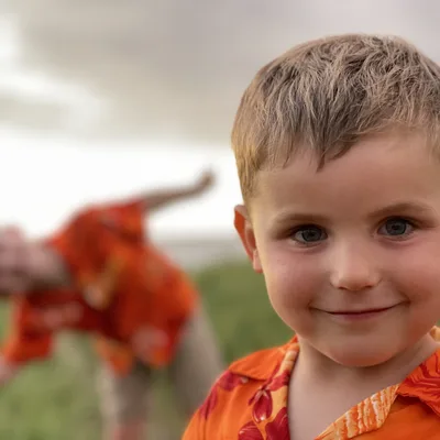 A young boy in an orange floral shirt smiles at the camera in a close-up portrait, while another child in matching attire is blurred in the background, posing playfully in an outdoor grassy field under an overcast sky.
