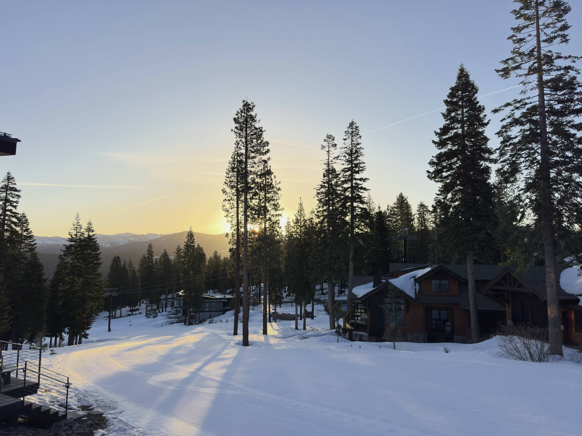 A snow-covered ski run winds past pine trees and wooden mountain cabins during a bright sunset. Long shadows stretch across the groomed snow, with distant mountains visible under a clear evening sky.