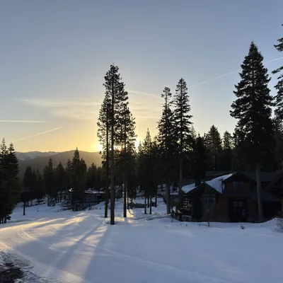 A snow-covered ski run winds past pine trees and wooden mountain cabins during a bright sunset. Long shadows stretch across the groomed snow, with distant mountains visible under a clear evening sky.