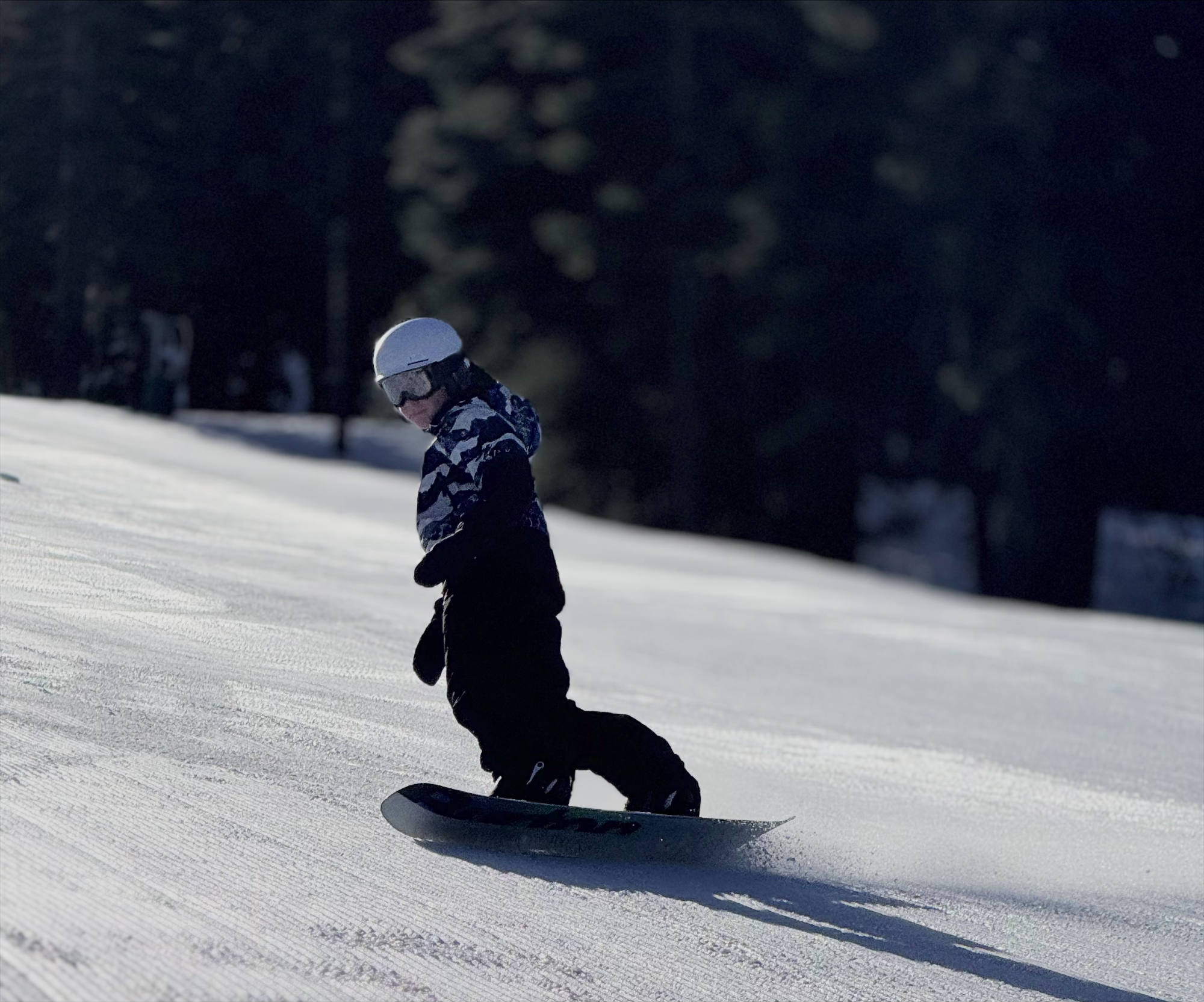 A person wearing a helmet, goggles, and winter gear is snowboarding down a groomed, snowy slope. The background features a dark, shadowed forest of evergreen trees.