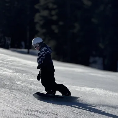 A person wearing a helmet, goggles, and winter gear is snowboarding down a groomed, snowy slope. The background features a dark, shadowed forest of evergreen trees.