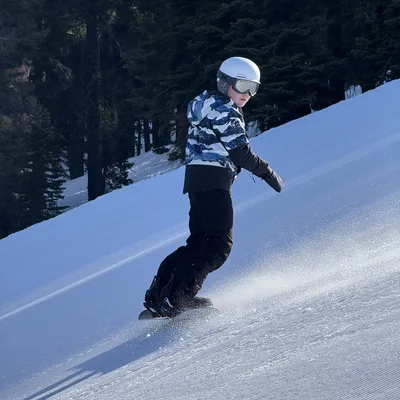 A person wearing a white helmet, goggles, and winter gear is snowboarding down a groomed, snowy slope. The background features a dense forest of evergreen trees under bright daylight.