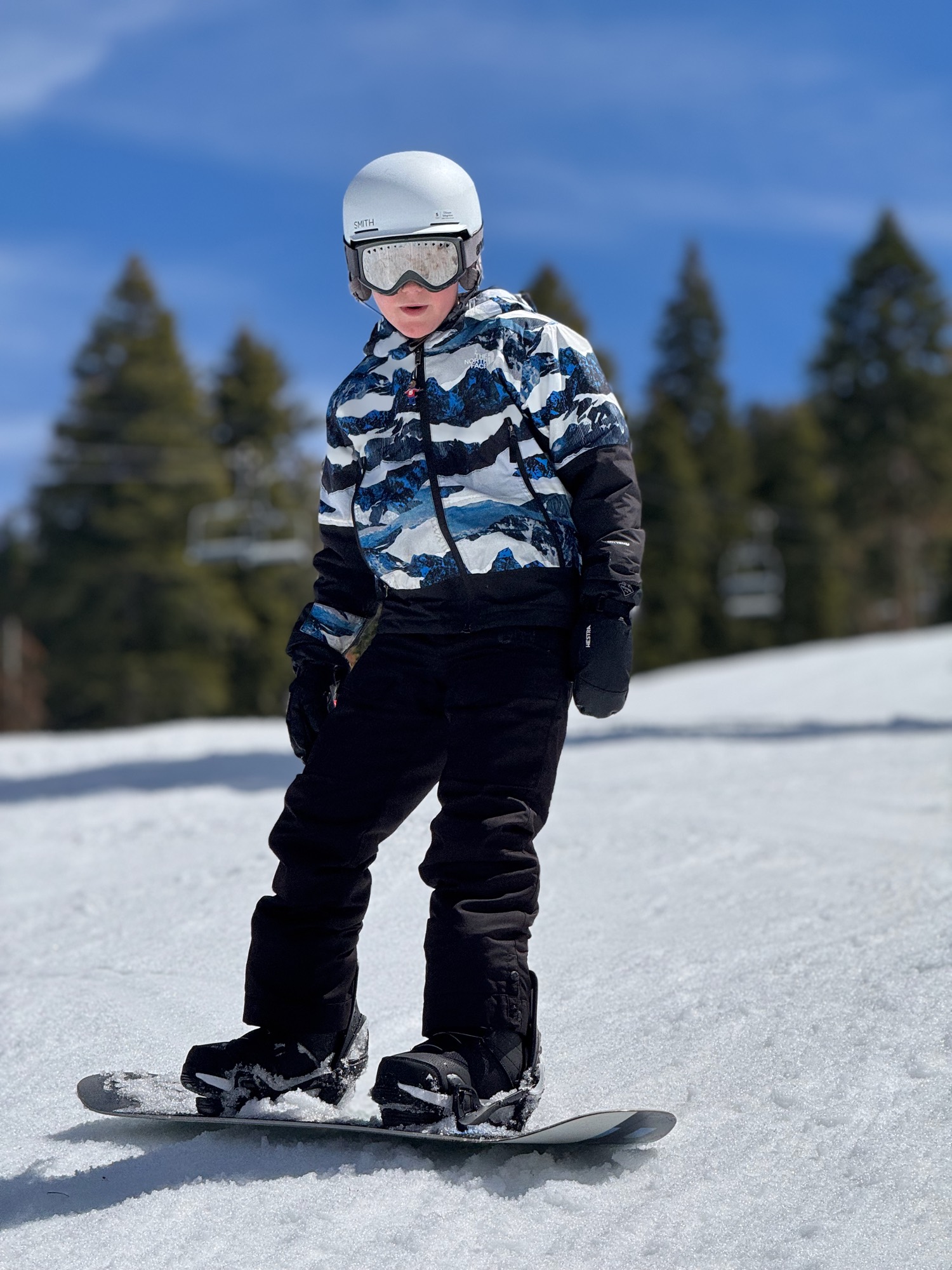 A young person wearing a helmet, goggles, and winter gear stands on a snowboard on a snowy slope. The background features evergreen trees and a clear blue sky on a sunny day.