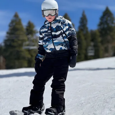 A young person wearing a helmet, goggles, and winter gear stands on a snowboard on a snowy slope. The background features evergreen trees and a clear blue sky on a sunny day.