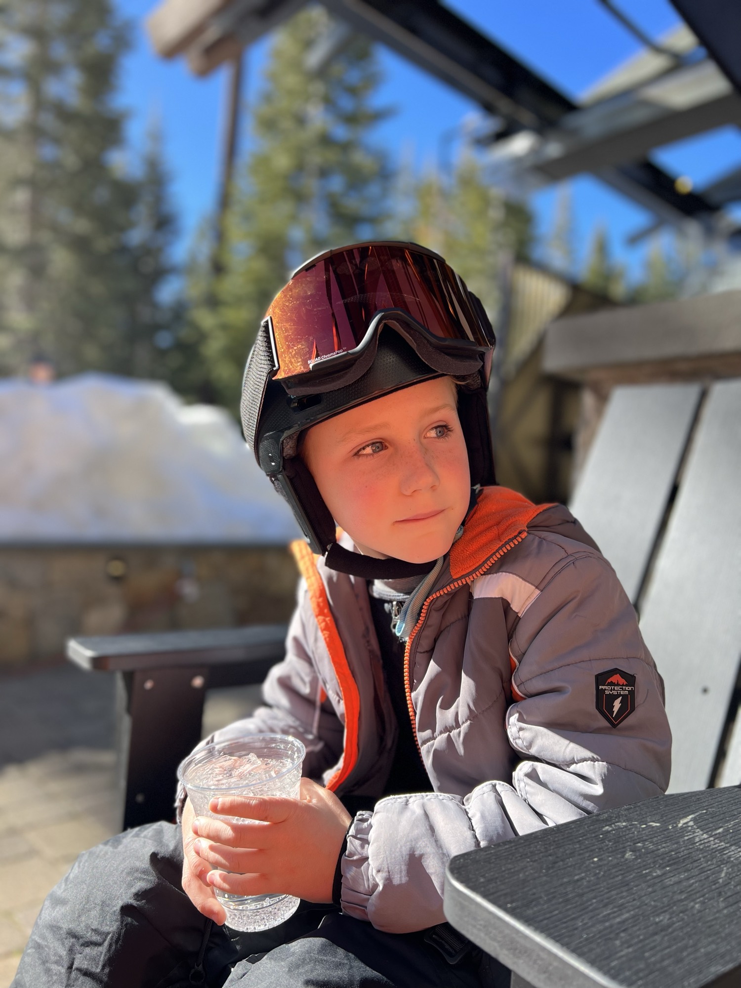 A young boy wearing a ski helmet, goggles, and winter jacket sits outdoors on a sunny day holding a plastic cup with a cold drink. He is seated at a table on a patio, with snow and pine trees visible in the background.