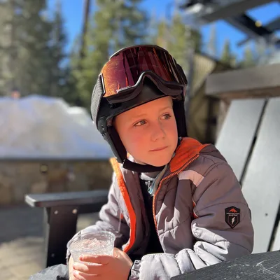 A young boy wearing a ski helmet, goggles, and winter jacket sits outdoors on a sunny day holding a plastic cup with a cold drink. He is seated at a table on a patio, with snow and pine trees visible in the background.