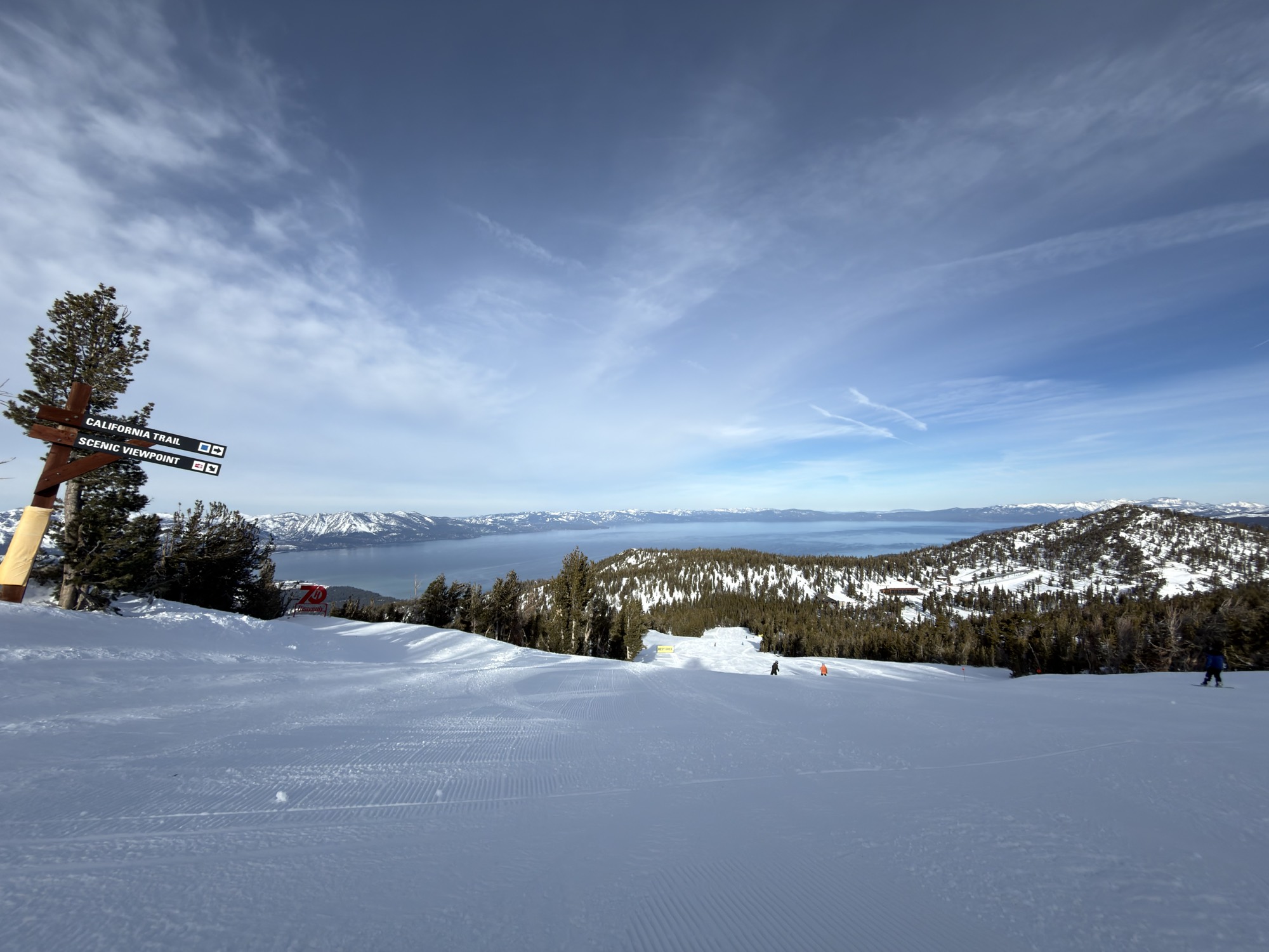 A wide shot of a snowy ski slope at a resort, featuring a wooden signpost pointing toward "California Trail" and "Scenic Viewpoint" on the left. In the background, a vast blue lake is surrounded by snow-dusted mountains under a partly cloudy sky, with a few distant skiers visible on the run.