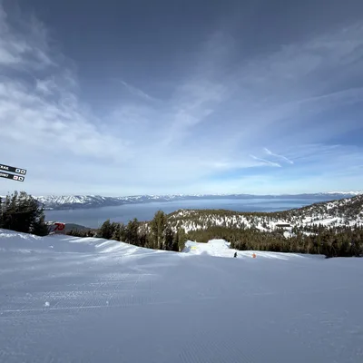 A wide shot of a snowy ski slope at a resort, featuring a wooden signpost pointing toward "California Trail" and "Scenic Viewpoint" on the left. In the background, a vast blue lake is surrounded by snow-dusted mountains under a partly cloudy sky, with a few distant skiers visible on the run.