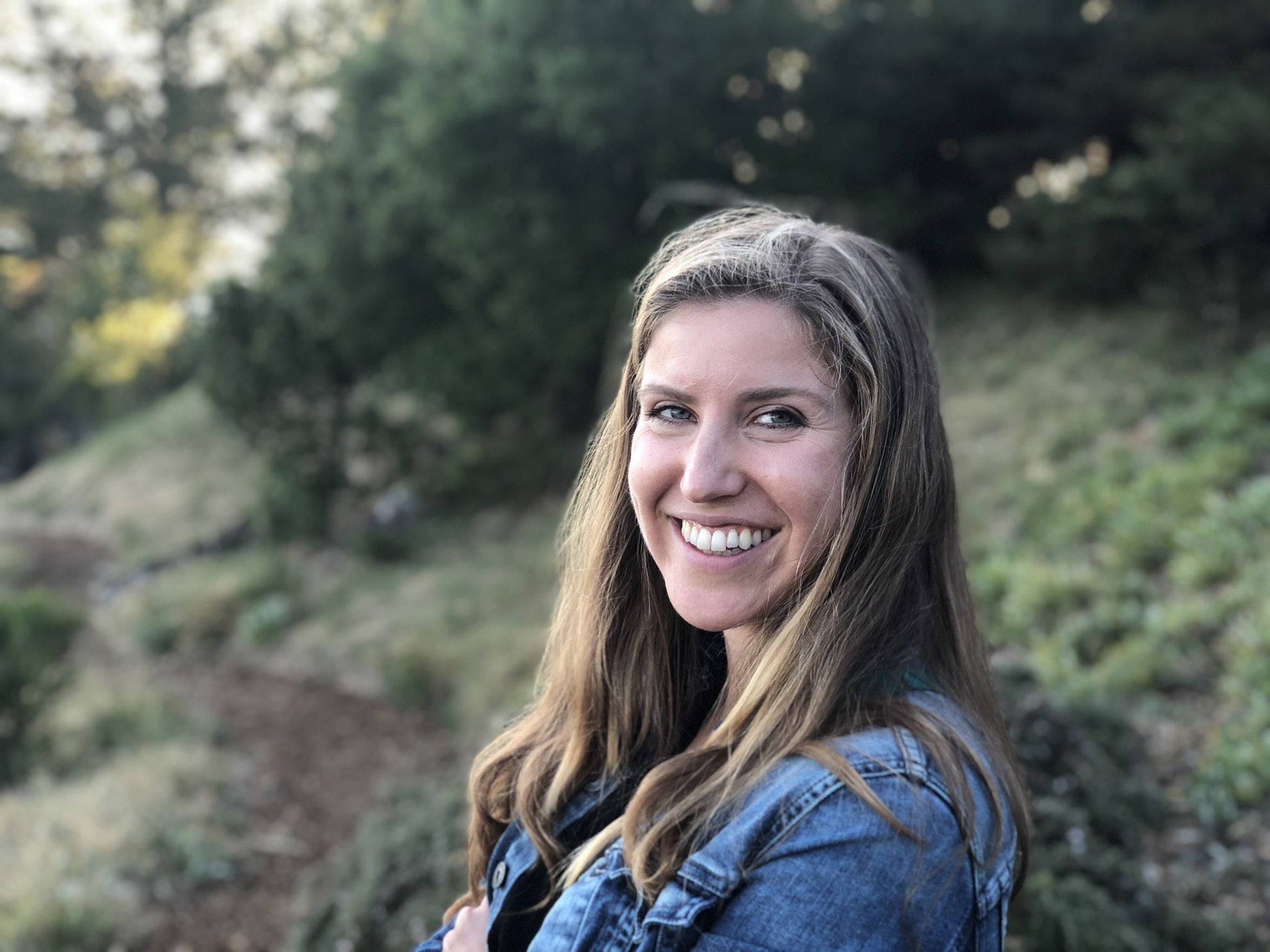 An outdoor portrait of a smiling woman with long brown hair wearing a denim jacket. She is standing on a grassy hillside with trees in the background.