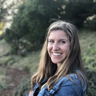 An outdoor portrait of a smiling woman with long brown hair wearing a denim jacket. She is standing on a grassy hillside with trees in the background.