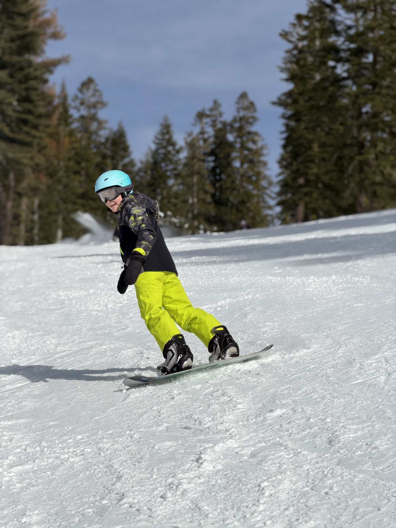 A young person wearing a blue helmet, goggles, and bright yellow pants is snowboarding down a snowy slope. The setting is an outdoor ski resort with pine trees lining the background under a clear blue sky.