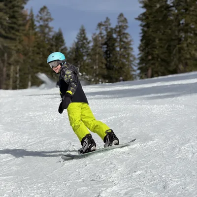 A young person wearing a blue helmet, goggles, and bright yellow pants is snowboarding down a snowy slope. The setting is an outdoor ski resort with pine trees lining the background under a clear blue sky.