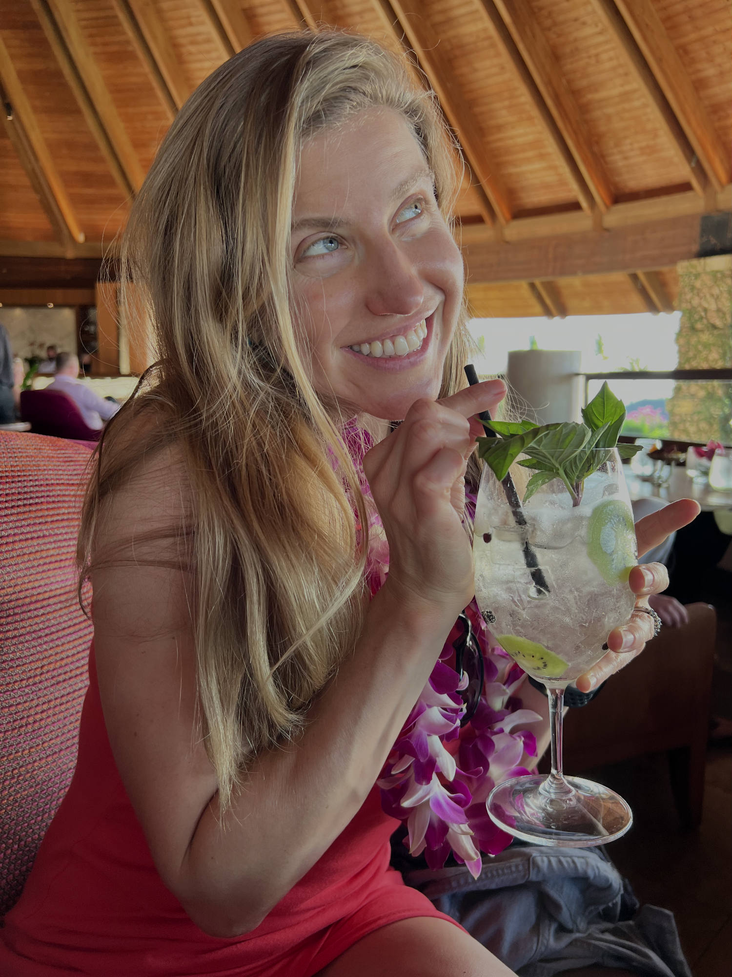 A smiling woman wearing a purple flower lei sits in an indoor restaurant with a wooden ceiling, holding a large cocktail garnished with basil and kiwi. She looks upward and away from the camera while holding the straw of her drink.