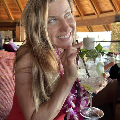 A smiling woman wearing a purple flower lei sits in an indoor restaurant with a wooden ceiling, holding a large cocktail garnished with basil and kiwi. She looks upward and away from the camera while holding the straw of her drink.