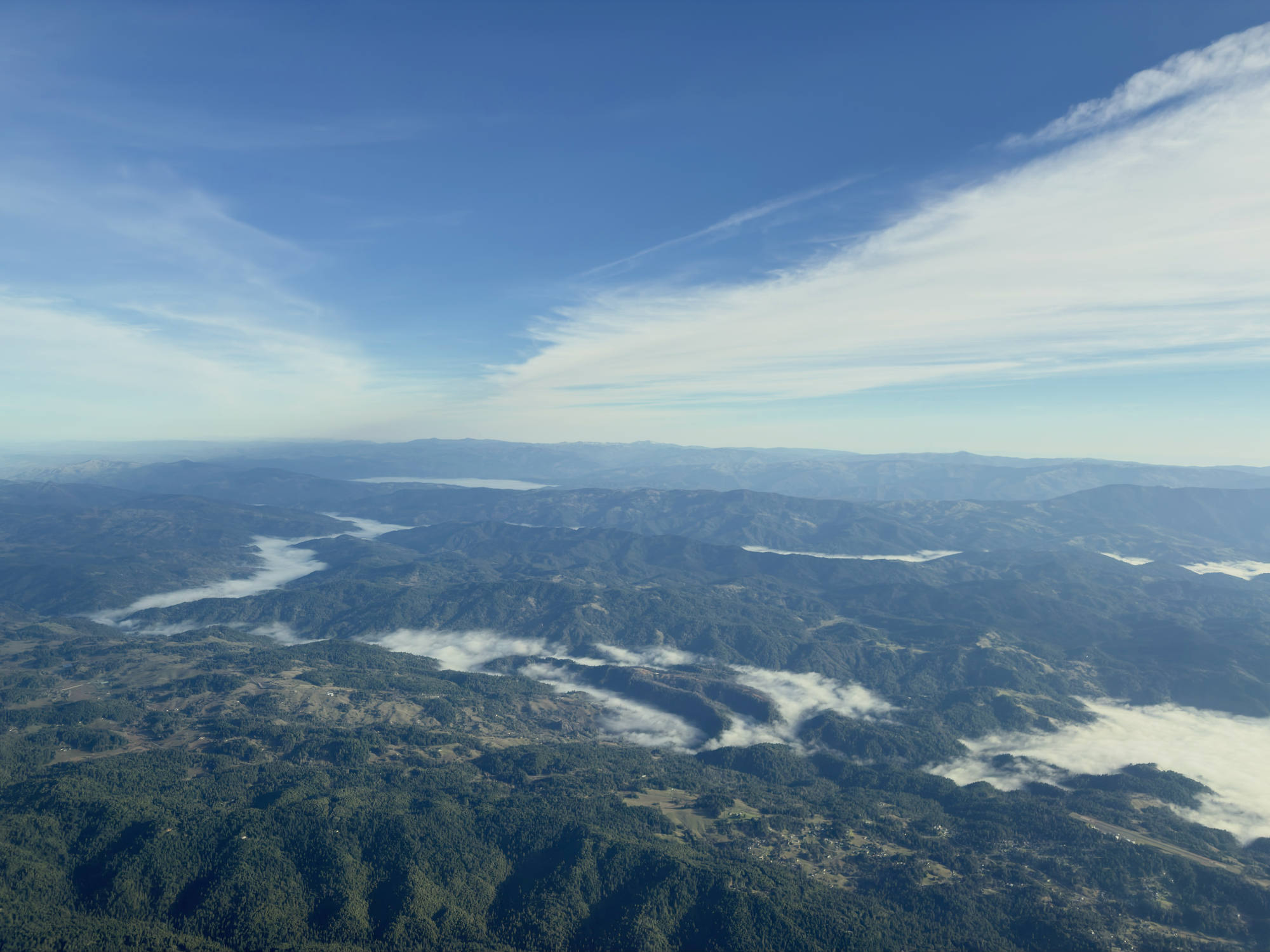 An aerial view captures a vast, mountainous landscape covered in green forests, with low-lying clouds and fog settling in the valleys. Above, a bright blue sky is filled with wispy, white cloud formations stretching across the horizon.