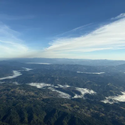 An aerial view captures a vast, mountainous landscape covered in green forests, with low-lying clouds and fog settling in the valleys. Above, a bright blue sky is filled with wispy, white cloud formations stretching across the horizon.