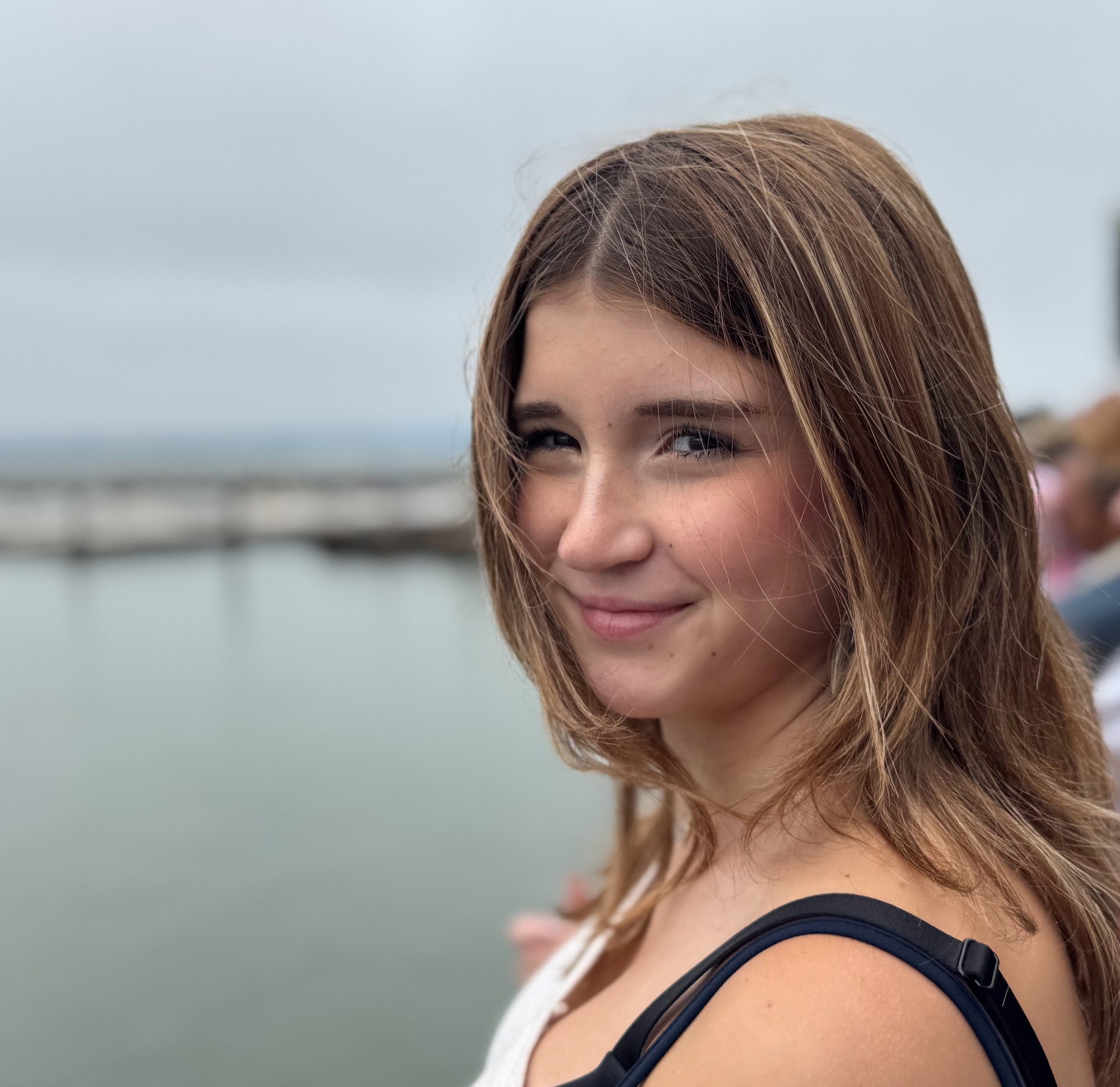 A close-up portrait shows a smiling young woman with light brown hair looking toward the camera. She is outdoors near a body of water on an overcast day, with a pier visible in the blurred background.