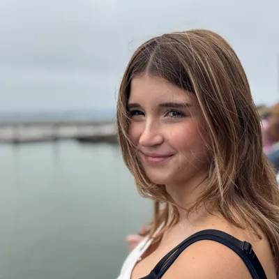 A close-up portrait shows a smiling young woman with light brown hair looking toward the camera. She is outdoors near a body of water on an overcast day, with a pier visible in the blurred background.