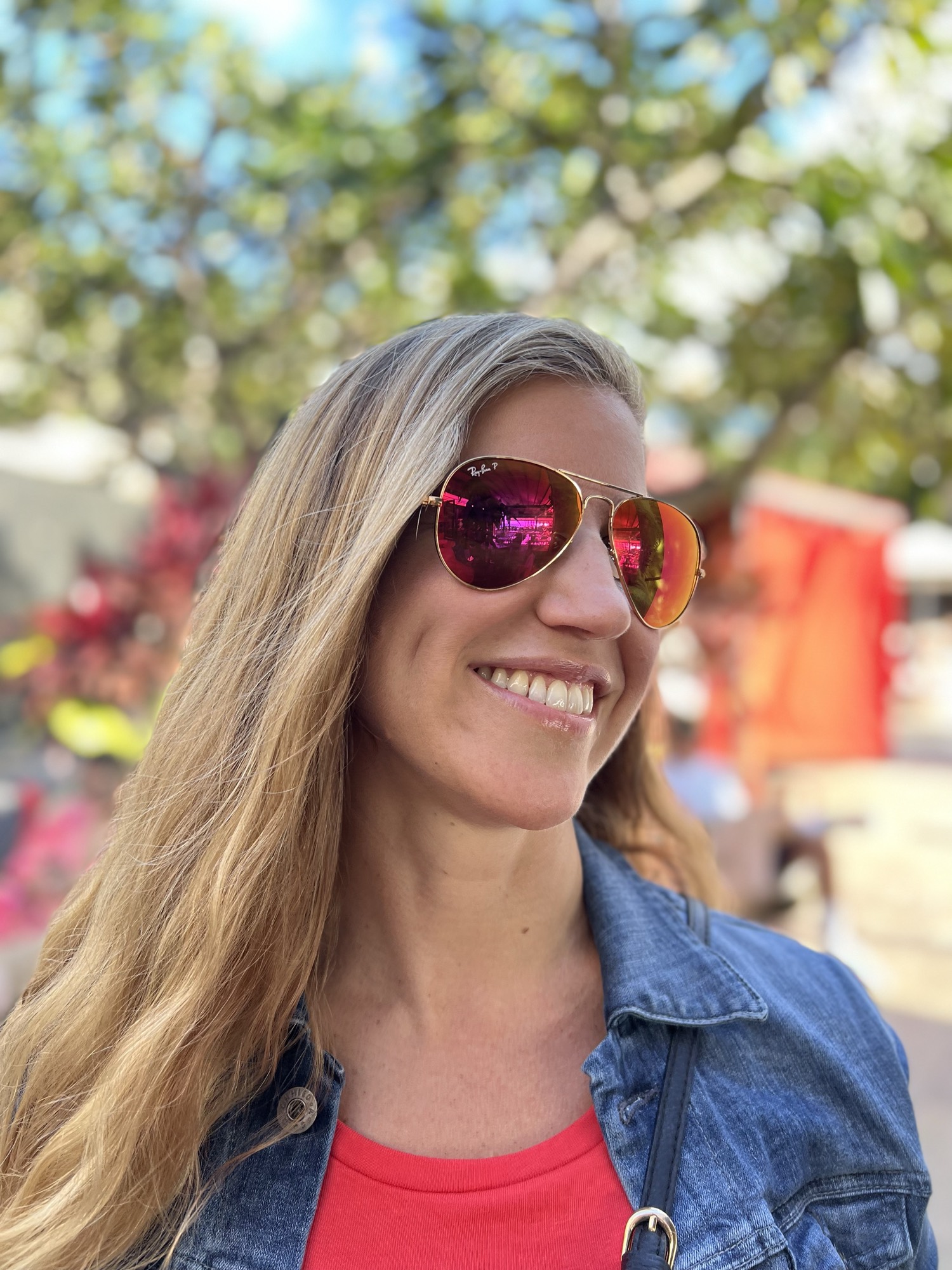 A close-up portrait shows a smiling woman with long blonde hair wearing a denim jacket and pink-tinted aviator sunglasses. She is outdoors on a sunny day with trees and a blurred background behind her.