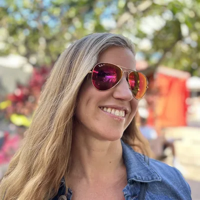 A close-up portrait shows a smiling woman with long blonde hair wearing a denim jacket and pink-tinted aviator sunglasses. She is outdoors on a sunny day with trees and a blurred background behind her.