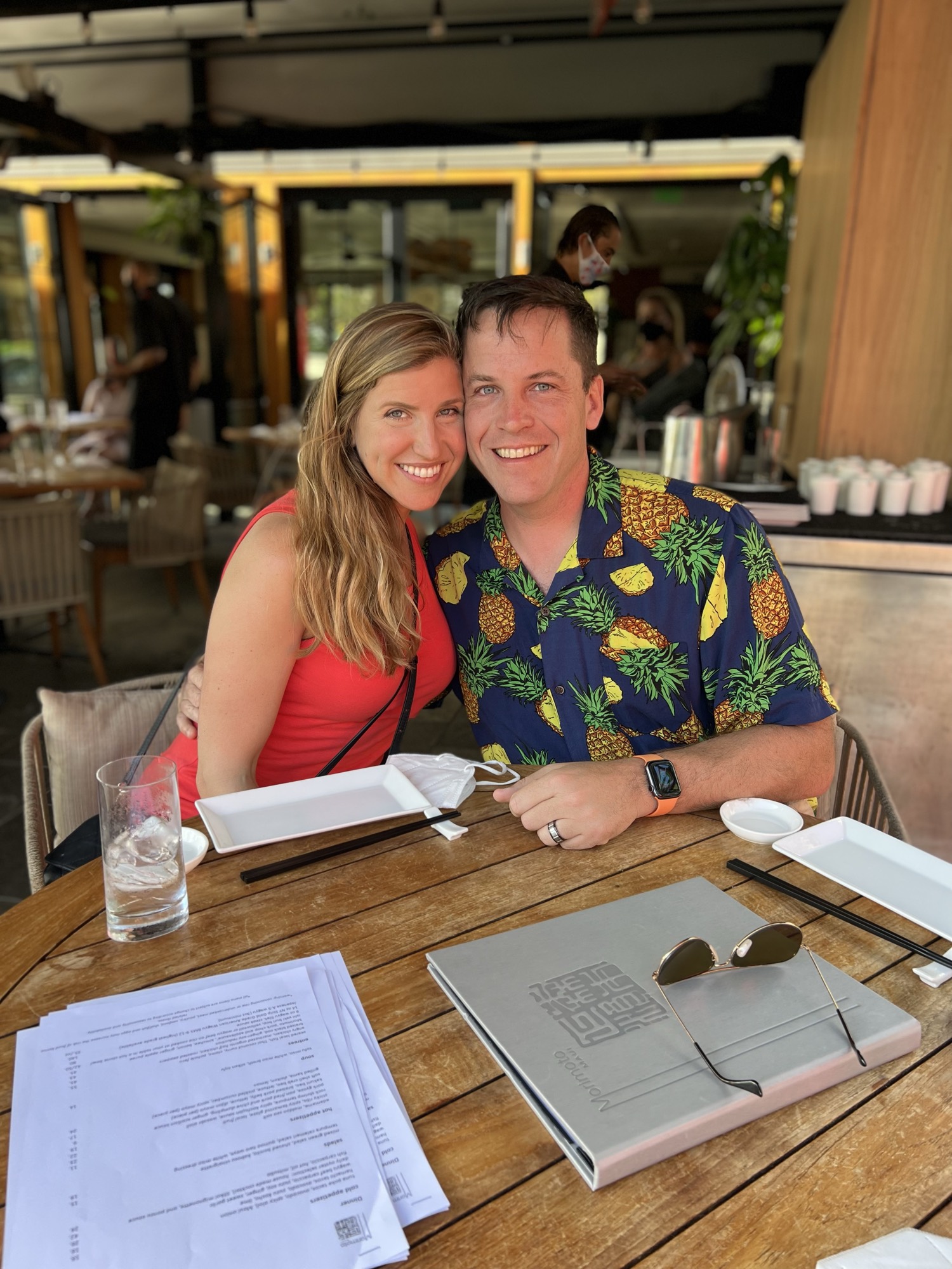 A smiling man in a pineapple-print shirt and a woman in a red top pose together at a wooden table in an outdoor restaurant. The table is set with menus, plates, chopsticks, a glass of water, and sunglasses.