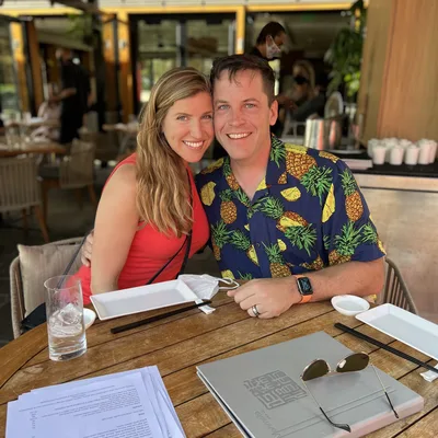 A smiling man in a pineapple-print shirt and a woman in a red top pose together at a wooden table in an outdoor restaurant. The table is set with menus, plates, chopsticks, a glass of water, and sunglasses.