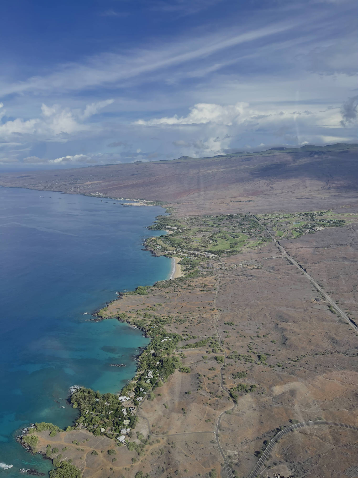 An aerial photograph captures a coastline with turquoise water, sandy beaches, and rocky shores bordering a dry, arid landscape. A long road runs parallel to the coast, leading toward a green golf course and resort area nestled against the volcanic slopes under a partly cloudy blue sky.
