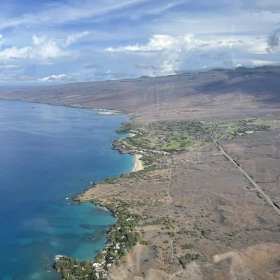 An aerial photograph captures a coastline with turquoise water, sandy beaches, and rocky shores bordering a dry, arid landscape. A long road runs parallel to the coast, leading toward a green golf course and resort area nestled against the volcanic slopes under a partly cloudy blue sky.