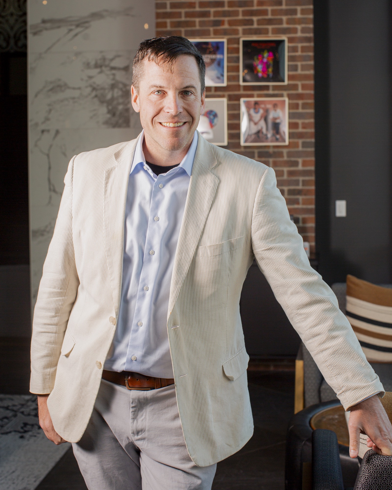 A smiling man in a light-colored blazer and blue button-down shirt stands indoors, posing for a portrait. He is positioned in a modern room featuring a brick accent wall with framed pictures and a marble-patterned wall in the background.
