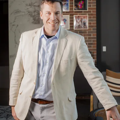 A smiling man in a light-colored blazer and blue button-down shirt stands indoors, posing for a portrait. He is positioned in a modern room featuring a brick accent wall with framed pictures and a marble-patterned wall in the background.