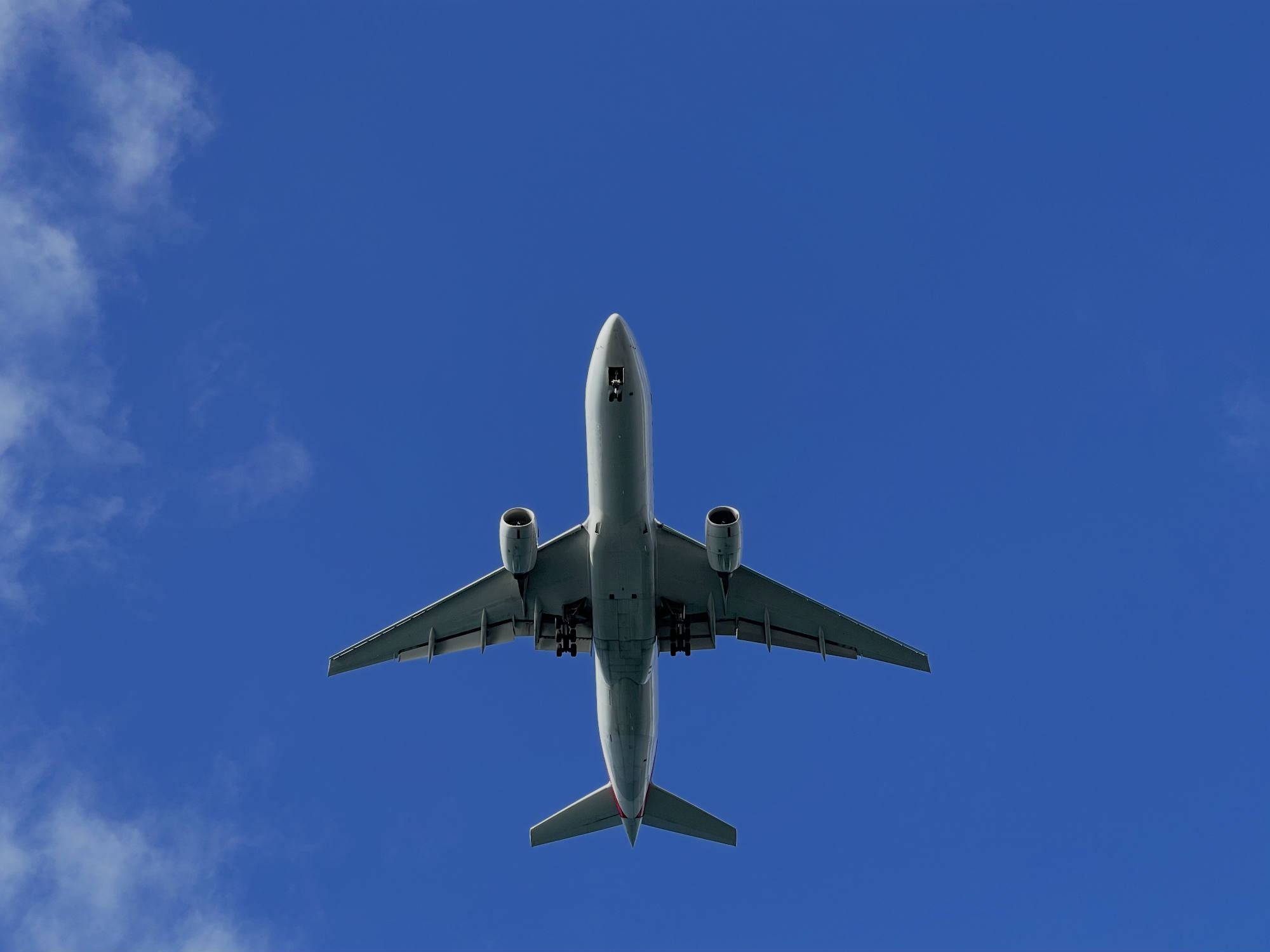 A low-angle shot captures a commercial airplane flying overhead with its landing gear deployed. The aircraft is centered against a bright blue sky with light, wispy clouds on the left.