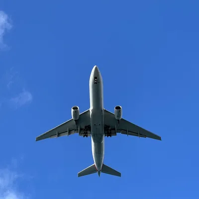 A low-angle shot captures a commercial airplane flying overhead with its landing gear deployed. The aircraft is centered against a bright blue sky with light, wispy clouds on the left.