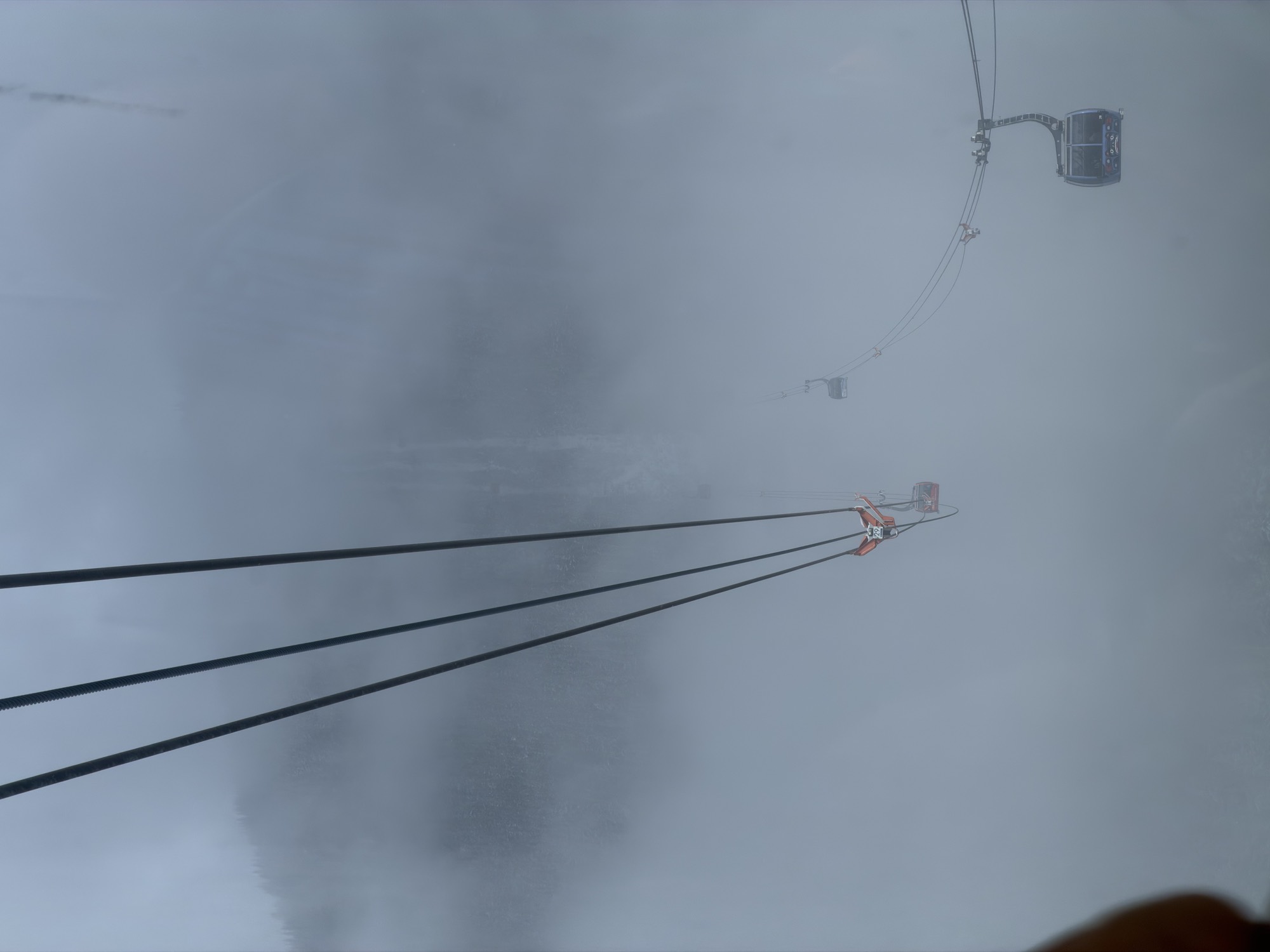 This high-angle shot captures cable cars traveling along suspension lines through a dense, foggy mountain landscape. The thick mist obscures the surrounding forest and terrain, creating a dramatic, atmospheric view of the transit system.
